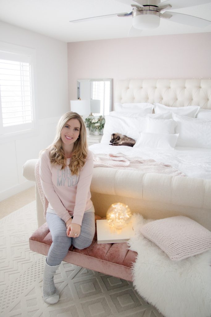 Hygge bedroom: Woman sitting in cozy pink and beige bedroom wearing pink Old Navy Cheers My Dear sweatshirt