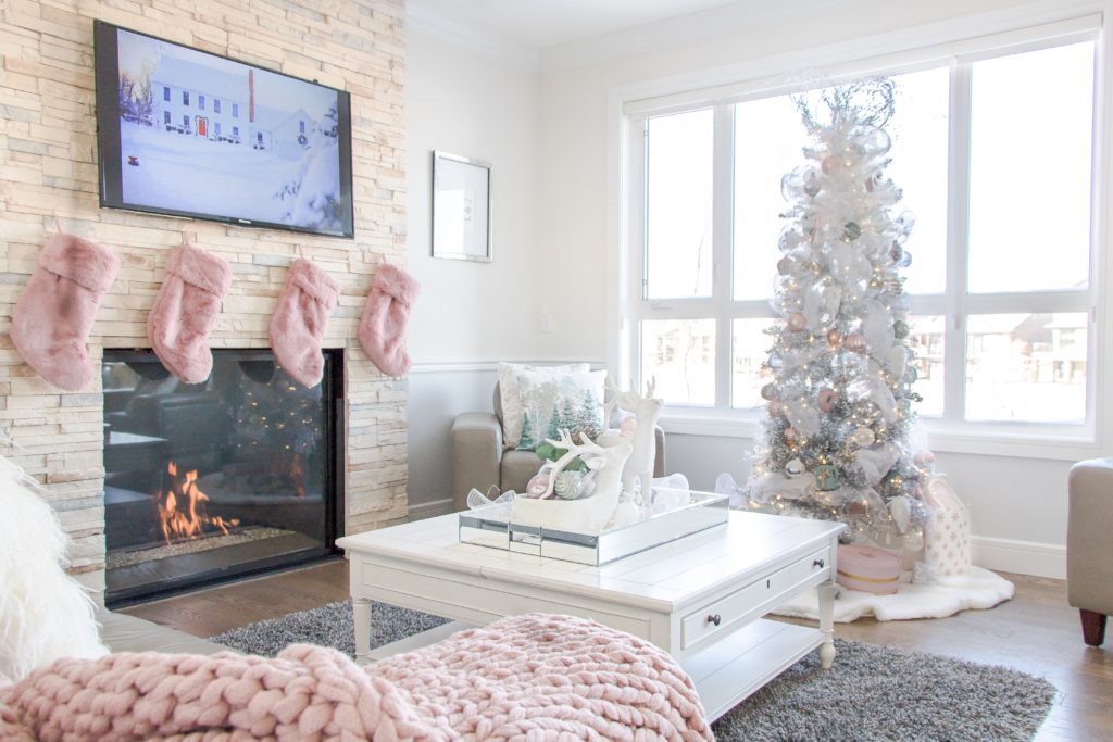 Christmas living room decor with pink fur stockings over a roaring fireplace, white and pastel pink Christmas coffee table decor, pink chunky knit blanket, ombre white and silver Christmas tree and pastel Christmas decor