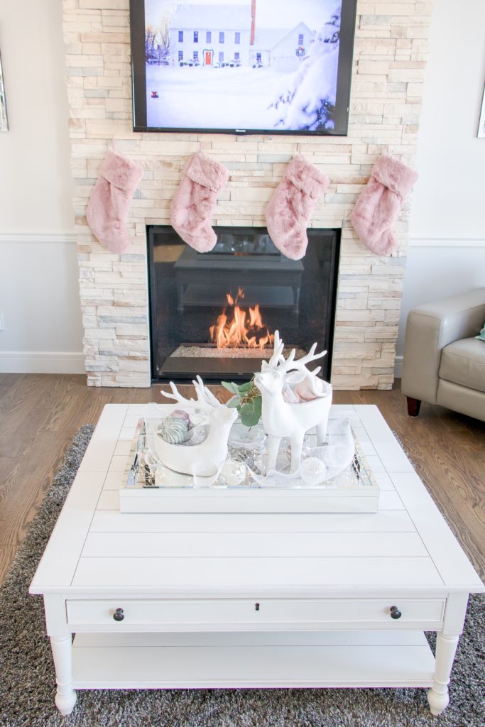 Christmas living room decor with pink fur stockings over a roaring fireplace, white and pastel pink Christmas coffee table decor, pink chunky knit blanket, ombre white and silver Christmas tree and pastel Christmas decor