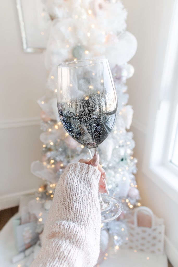 Silver shimmering wine glass with a white Christmas tree with pastel toned ornaments in the background, glamorous Christmas decorating ideas