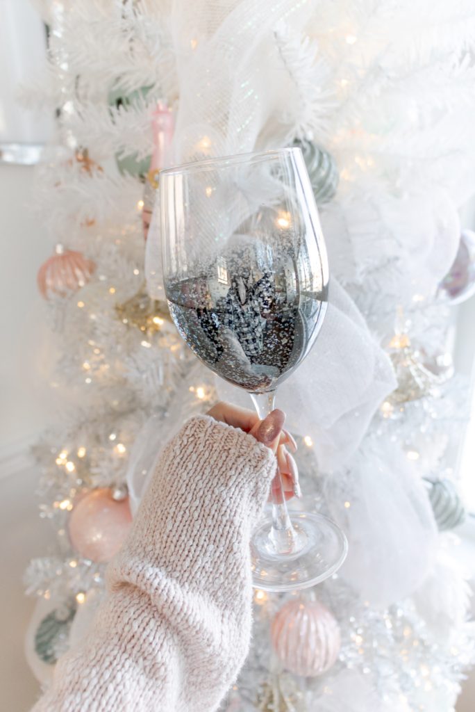 Silver shimmering wine glass with a white Christmas tree with pastel toned ornaments in the background, glamorous Christmas decorating ideas