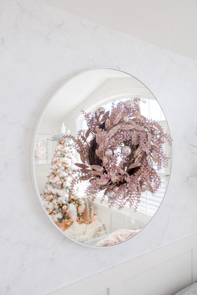 Pink glitter wreath hung on a circular living room mirror with the reflection of a white fluffy Christmas tree with pastel tones