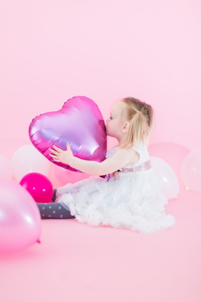 Toddler Valentine's Day photo in white rosette dress from Monsoon with heart-shaped balloons - Cute Valentine's Day outfits for little girls, little boys and women - Valentine's Day outfit inpso