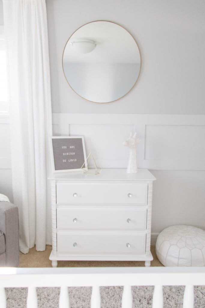 White gender-neutral nursery dresser with gold circular mirror and white board and batten wainscoting