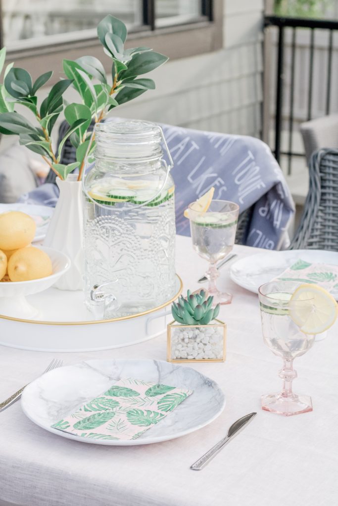 Closeup of easy outdoor table setting featuring marble-look outdoor plates, faux greenery, pink goblets and palm print napkins