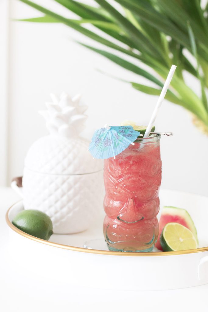 Frozen Water-Yellin’ Watermelon Tiki Drink on a white tray with fresh limes, watermelon and a ceramic pineapple with palm leaves in the background