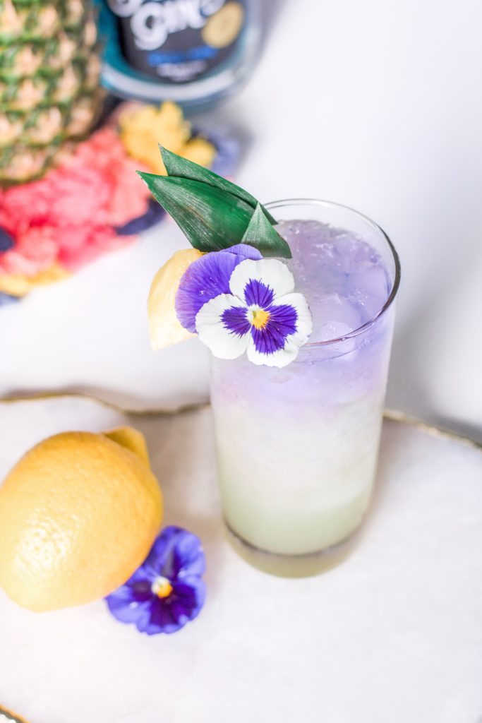 Closeup of Layered Gin Pineapple Cocktail, garnished with purple pansy, a fresh pineapple chunk and pineapple leaves