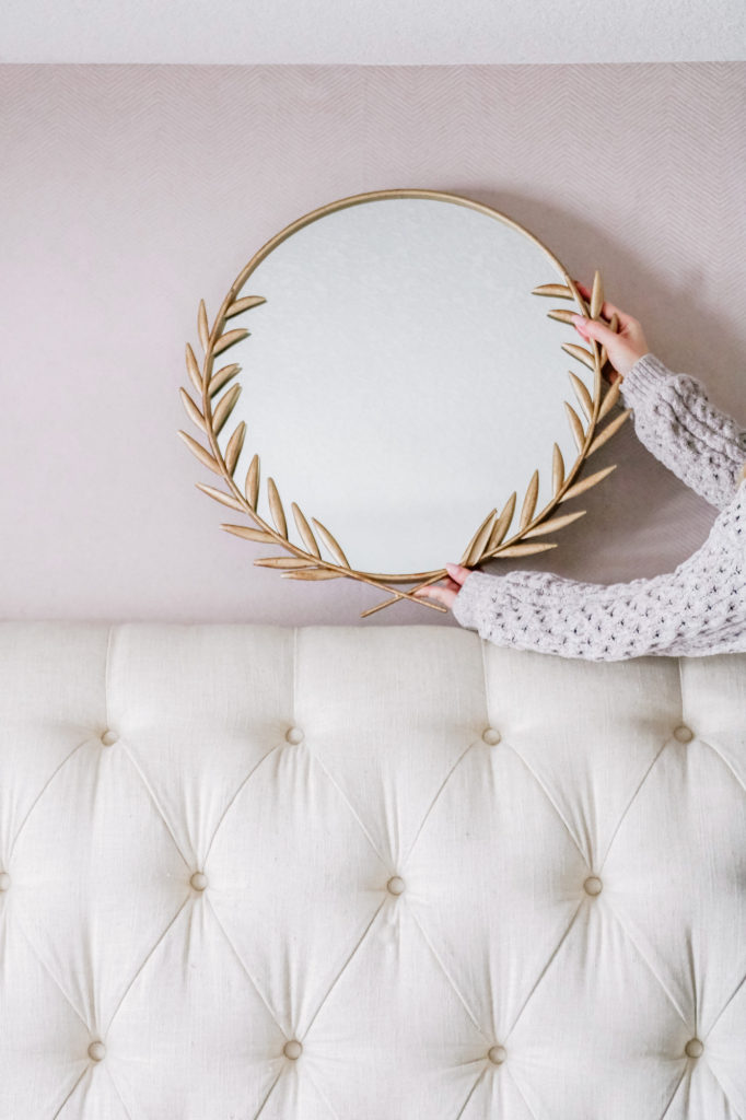 Gold wreath mirror overtop tufted bed with pink wallpaper